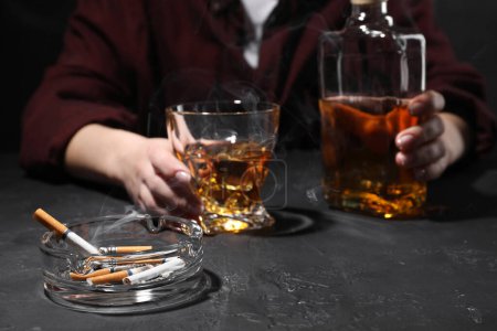 Adicción al alcohol. Mujer con whisky y cigarrillos humeantes en mesa de textura oscura, enfoque selectivo