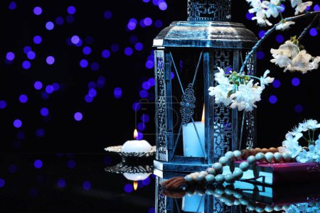 Arabic lantern, Quran, misbaha, burning candle and flowers on mirror surface against blurred lights at night. Space for text