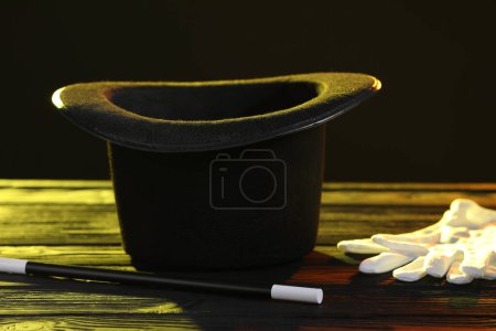 Magician's hat, wand and gloves on wooden table against black background