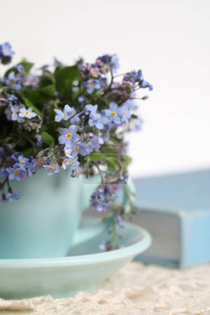 Beautiful forget-me-not flowers in cup, book and crochet tablecloth on table against white background, closeup