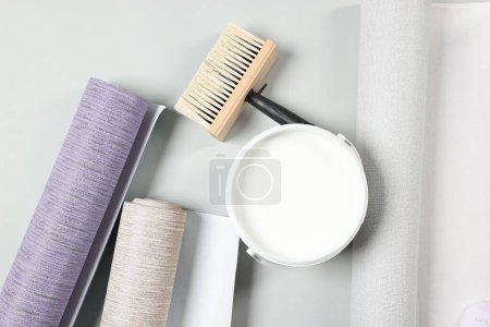 Wallpaper rolls, brush and bucket of glue on light grey background, flat lay