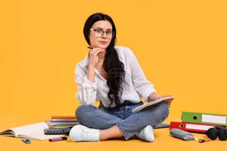 Estudiante con cuaderno sentado entre libros y papelería sobre fondo amarillo