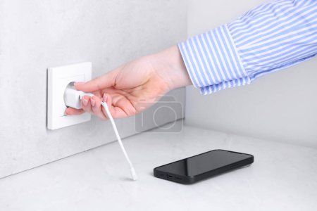 Woman plugging smartphone into power socket at white table indoors, closeup