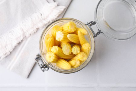 Maïs bébé jaune frais savoureux en pot en verre sur table carrelée blanche, vue sur le dessus