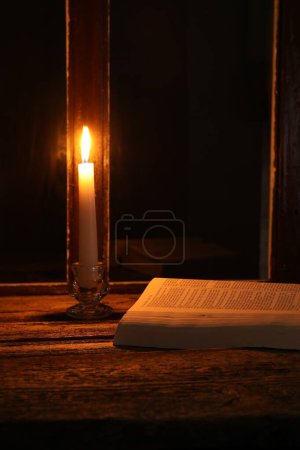 Burning candle and Bible on wooden table near window at night