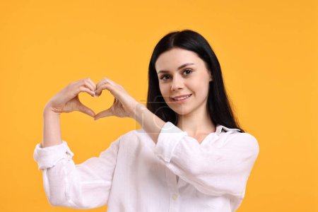 Lächelnde Frau zeigt Herz-Geste mit Händen auf orangefarbenem Hintergrund