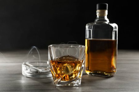 Adicción al alcohol. Whisky en vidrio, botella, cigarrillos y cenicero sobre mesa de madera