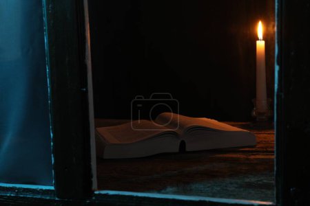 Burning candle and Bible on wooden table at night, view through window