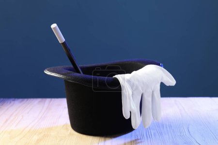 Magician's hat, wand and gloves on wooden table against blue background