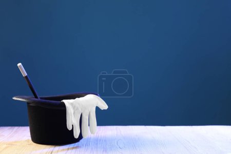 Magician's hat, wand and gloves on wooden table against blue background, space for text