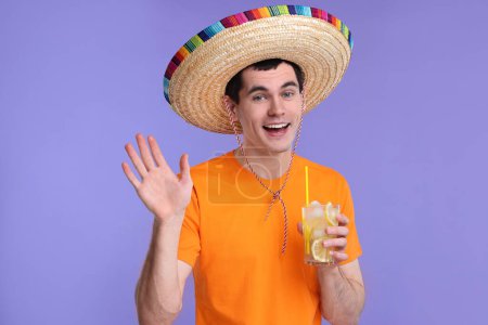 Hombre joven en sombrero mexicano sombrero con cóctel saludando sobre fondo violeta