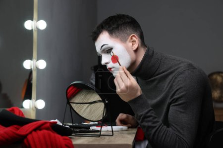 Young man applying mime makeup near mirror in dressing room