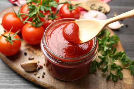 Photo for Jar and spoon with tasty ketchup, fresh tomatoes, parsley and spices on grey wooden table, closeup - Royalty Free Image