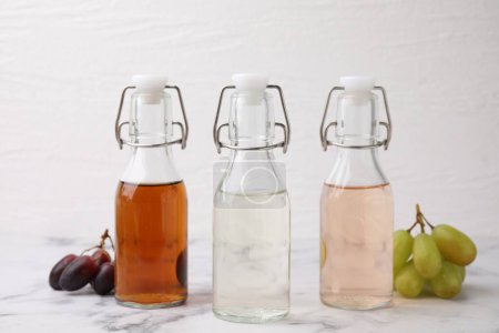 Different types of vinegar in bottles and grapes on light marble table, closeup