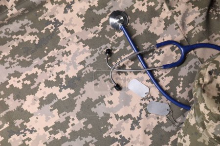Stethoscope, military ID tags and cap on camouflage fabric, flat lay. Space for text