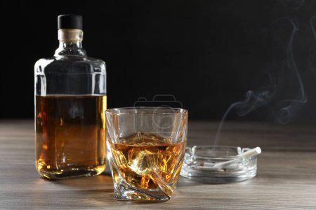 Téléchargez les photos : L'alcoolisme. Whisky en verre, bouteille, cigarettes et cendrier sur table en bois - en image libre de droit