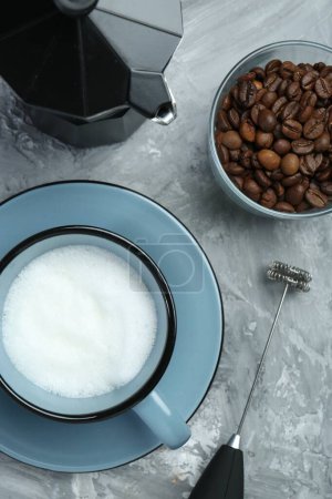 Mini mixer (milk frother), whipped milk in cup, coffee beans and moka pot on grey textured table, flat lay