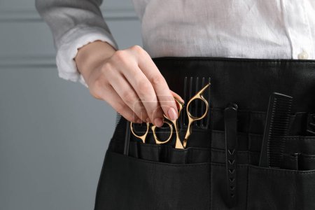 Hairstylist with professional tools in waist pouch near grey wall, closeup