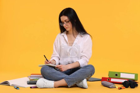Student writing in notebook among books and stationery on yellow background