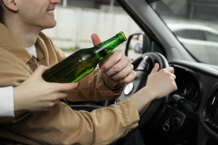 Téléchargez les photos : Un homme souriant prend une bouteille de bière en conduisant une voiture, gros plan. Ne pas boire et conduire concept - en image libre de droit