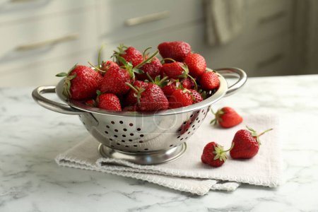Metal colander with fresh strawberries on white marble table in kitchen