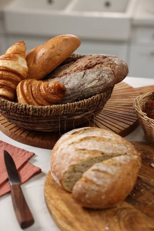 Wicker bread basket with freshly baked loaves and knife on white marble table in kitchen, closeup