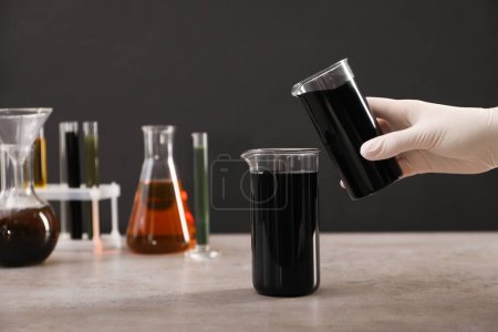 Photo for Woman pouring black crude oil into beaker at grey table against dark background, closeup - Royalty Free Image