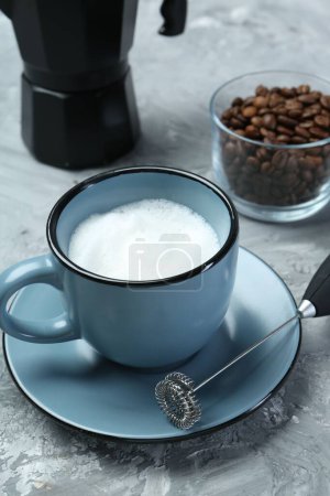 Mini batidora (espuma de leche), leche batida en taza y granos de café en mesa texturizada gris