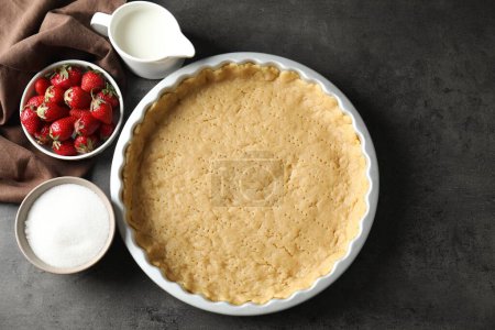 Making shortcrust pastry. Raw dough in baking dish, milk, sugar and strawberries on grey table, top view