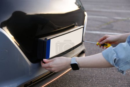 Woman with screwdriver installing vehicle registration plate to car outdoors, closeup