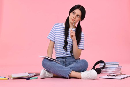 Student with notebook sitting among books and stationery on pink background