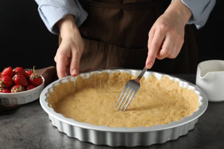 Shortcrust pastry. Woman making holes in raw dough with fork at grey table, closeup