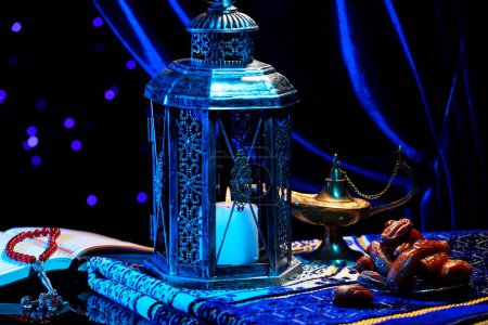 Arabic lantern, Quran, misbaha, Aladdin magic lamp, dates and folded prayer mat on mirror surface against blurred lights at night