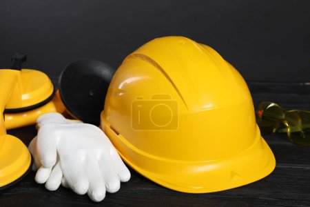 Hard hat, suction lifters, protective gloves and goggles on black wooden surface against gray background, closeup