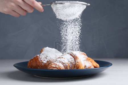 Woman with sieve sprinkling powdered sugar onto croissants at white wooden table, closeup