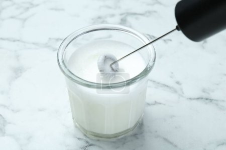 Batiendo la leche en el vaso con el mini mezclador (espuma de leche) en la mesa de mármol blanco, primer plano