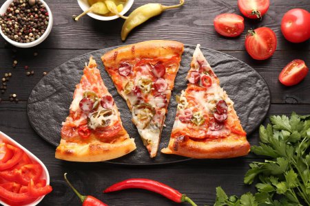 Slate board with pieces of delicious pizza Diablo among products on black wooden table, flat lay
