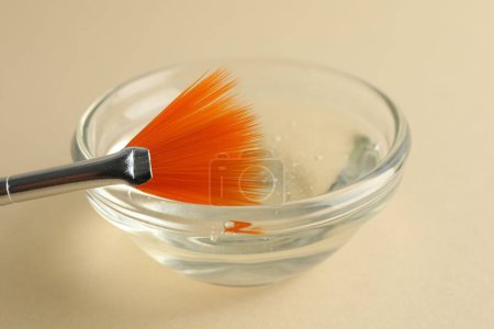 Chemical peel liquid in bowl and brush on beige background, closeup. Peeling procedure