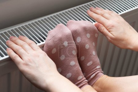Téléchargez les photos : A woman warms her feet and hands in socks on a radiator. Home heating. Low temperature and cold in the house. Photo closeup - en image libre de droit