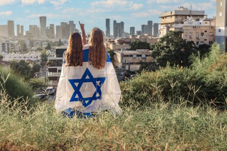 Photo for Two girls are wrapped up in the Israel flag and looking at the horizon. cityscape of Givatayim in the background. Yom Haatzmaut or Stand with Israel concept - Royalty Free Image