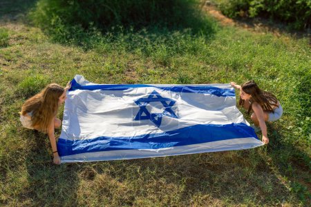 Photo for Two girls spread the flag of Israel as a tablecloth on the grass for a picnic. Yom Haatzmaut or Stand with Israel concept - Royalty Free Image