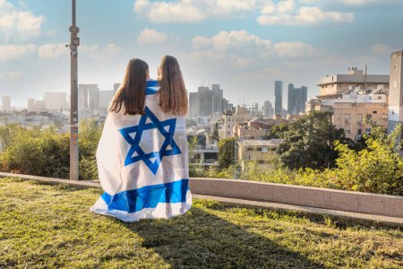 Two girls are wrapped up in the Israel flag and looking at the horizon. cityscape of Givatayim in the background. Yom Haatzmaut or Stand with Israel concept