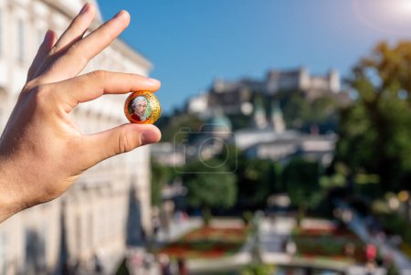 Salzburg, Austria, August 15 2022. A Mozart ball between forefinger and thumb of one hand: in the blurred background Mirabell Castle and Hohensalzburg Fort. Symbolic image.