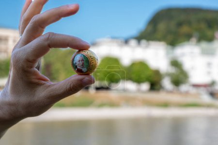 Salzburgo, Austria, 15 de agosto 2022. Una bola de Mozart entre el dedo índice y el pulgar de una mano: en el fondo borroso Castillo de Mirabell y el Fuerte de Hohensalzburg. Imagen simbólica.