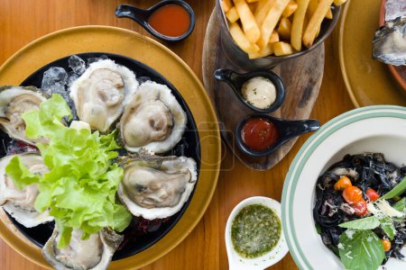 Téléchargez les photos : Un plateau d'huîtres crues bio fraîches sur glace, frites et fettuccini noir avec sauce à l'encre noire servi sur une table en bois. Vue du dessus - en image libre de droit