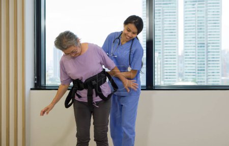 Une physiothérapeute en uniforme bleu aide une personne âgée à faire de l'exercice et à marcher avec sa ceinture de sécurité. Atmosphère dans le centre de santé communautaire.