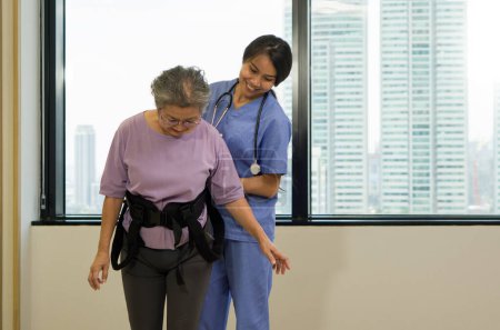 Téléchargez les photos : Une physiothérapeute en uniforme bleu aide une personne âgée à faire de l'exercice et à marcher avec sa ceinture de sécurité. Atmosphère dans le centre de santé communautaire. - en image libre de droit