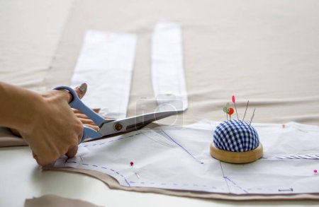 Foto de Primer plano dressmaker mano utilizar tijeras para cortar tela beige fina y patrón de papel de boceto. - Imagen libre de derechos
