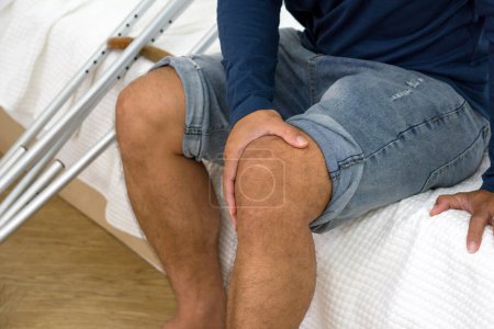 Injured man with painful knee resting on bed in an indoor setting, displaying discomfort. Crutches were placed beside him.