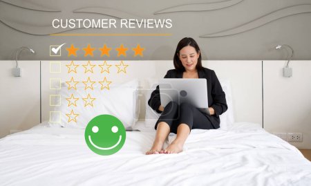 Foto de Mujer de negocios enfocada hojeando a través de comentarios de los clientes en su computadora portátil mientras se sienta cómodamente en una cama en un ambiente tranquilo y sereno. - Imagen libre de derechos